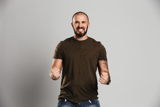 Photo of handsome tattooed guy with beard and mustache clenching his fists showing strength, isolated over gray wall