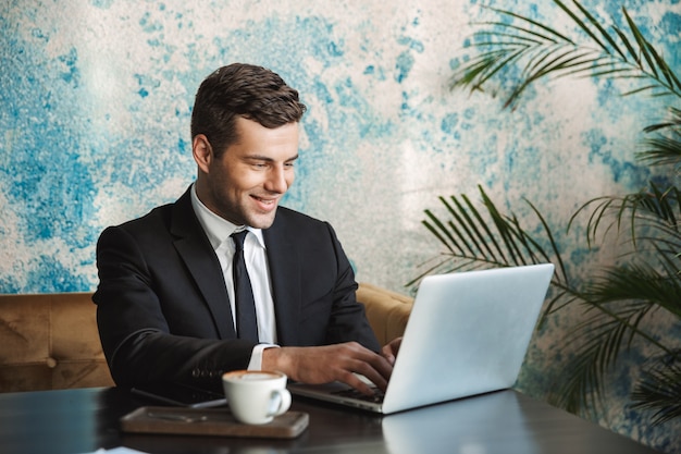 Photo of handsome young businessman in formal clothes indoors in cafe using laptop computer.