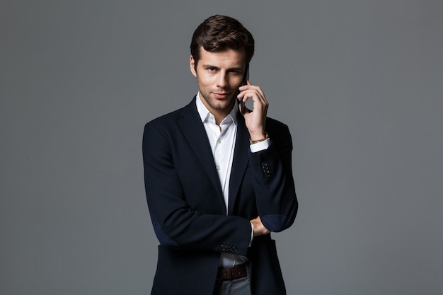 Photo of a handsome young concentrated business man isolated over grey wall wall talking by mobile phone.