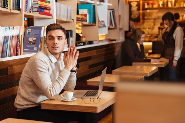 Photo of handsome young man sitting in cafe and chatting by laptop computer. Looking aside.