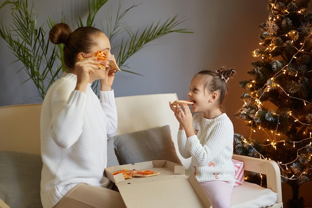 Foto di felice madre e figlia che indossano un maglione bianco seduti sul divano vicino all'albero di natale e mangiano pizza donna divertendosi e coprendosi gli occhi con fette di pizza