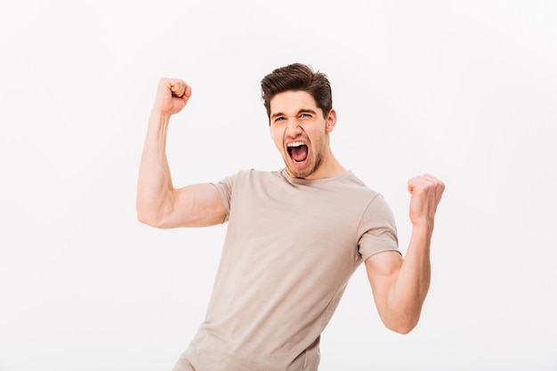 Photo of happy brunette guy rejoicing and clenching fists like winner or lucky person, isolated over white wall