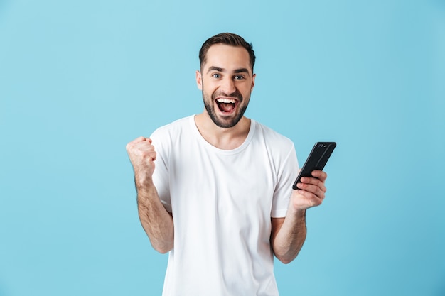 Photo of happy brunette man wearing basic t-shirt laughing and holding smartphone isolated over blue 