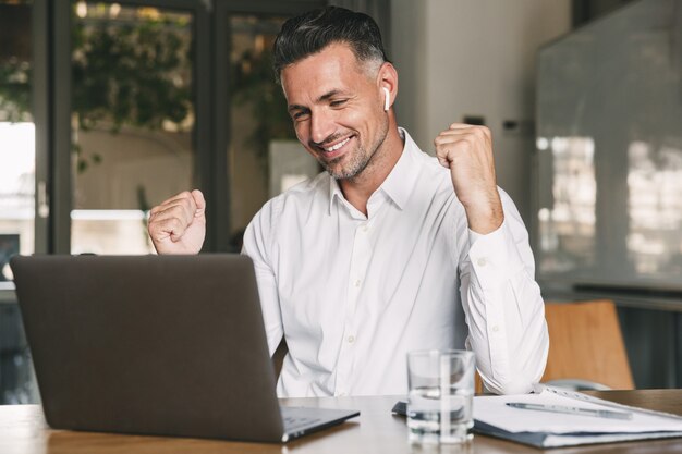 Photo of happy businessman 30s wearing white shirt and wireless earphones rejoicing and clenching fist, while working on laptop in office