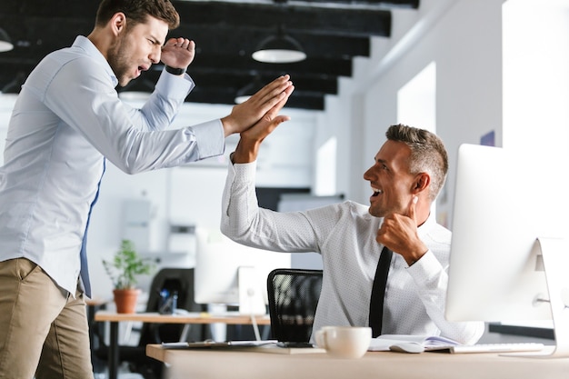 Photo photo of happy businessmen 30s in formal clothes giving high five together in office, during successful deal