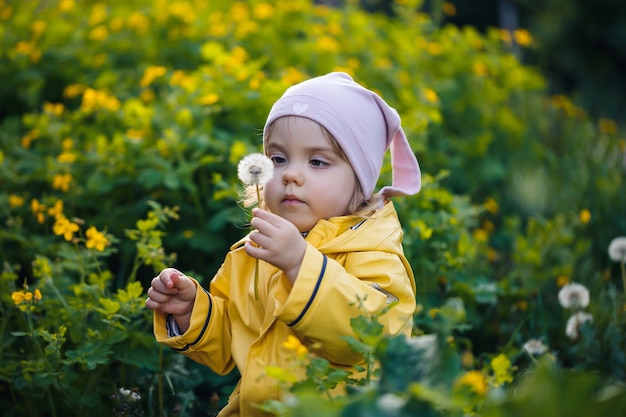 Foto di una bambina felice che indossa una giacca gialla e un cappello bianco che gioca in un prato, fiori circondati da un bambino ed erba, che gioca nel campo durante il tramonto. concetto di infanzia.