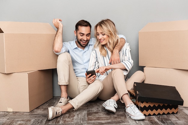 Photo of happy couple in casual clothing seating near cardboard boxes and looking at smartphone isolated over gray wall