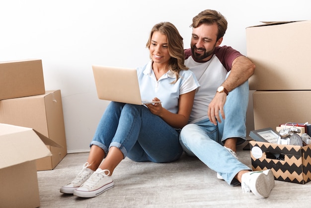 Photo of happy couple in casual clothing using laptop and hugging while sitting near cardboard boxes isolated over white wall