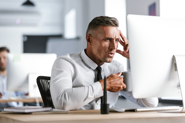 Foto di felice regista uomo 30s indossa camicia bianca e cravatta bere il tè dalla tazza, mentre era seduto al computer in ufficio