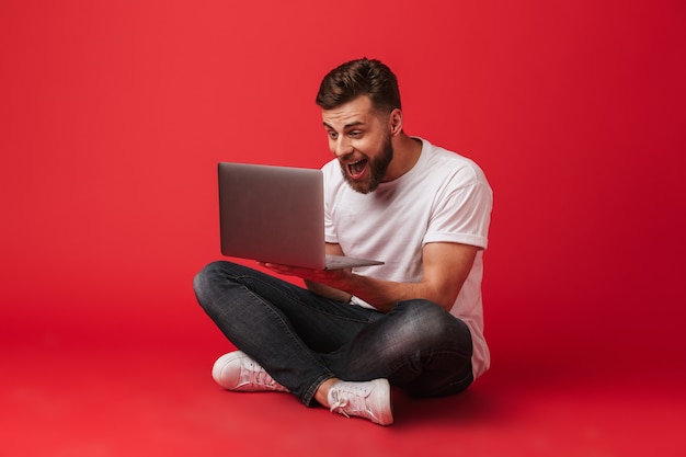 Photo of happy excited man in t-shirt and jeans sitting on floor and screaming like lucky guy or winner while looking on laptop, isolated over red background