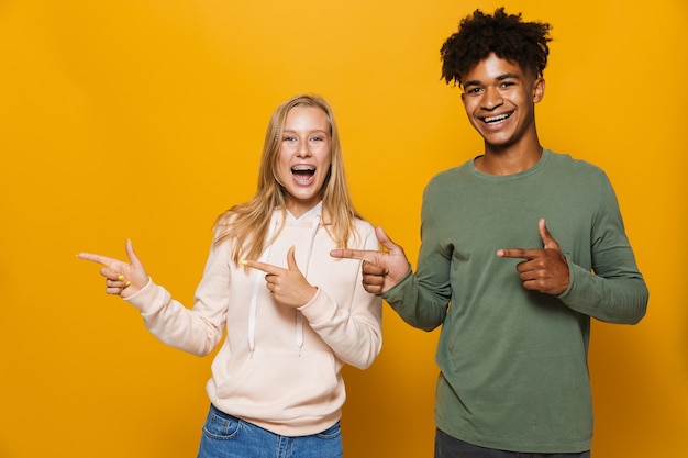 幸せな友人の男性と女性の写真16-18は、黄色の背景の上に分離された、コピースペースで笑って指を脇に向けて笑っている歯列矯正器を持っています