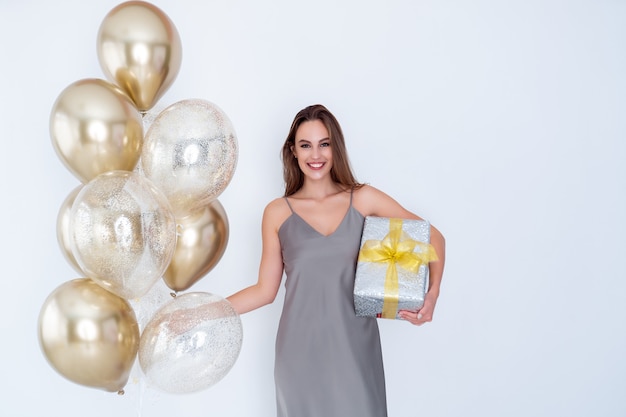 Photo of happy girl holds large wrapped giftbox and many air balloons came to party celebration