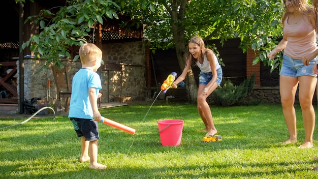 Photo of happy laughing family splashing water with water guns and garden hose at backyard. People playing and having fun on hot sunny summer day