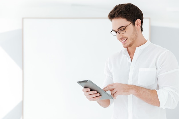 Photo of happy man wearing eyeglasses and dressed in white shirt using tablet computer near big board. Look at tablet.