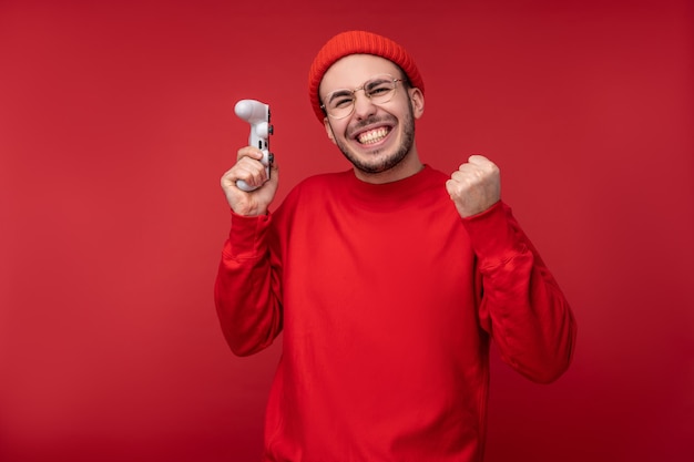 Photo of happy man wih beard in red clothing holds joystick playing game winner with surprise and success, isolated over red background.