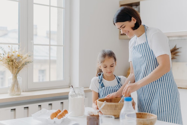 Photo of happy mum and child cook together at kitchen, wears\
aprons prepare something tasty, make food, whisk eggs with beater,\
bake house pastries at home. milk, eggs, chocolate, flour on\
table