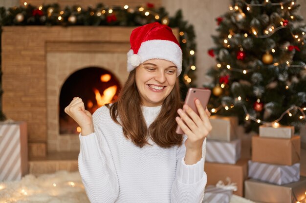 La foto della ragazza sorridente felice legge le notizie di natale sullo smartphone, alza i pugni mentre si siede in casa