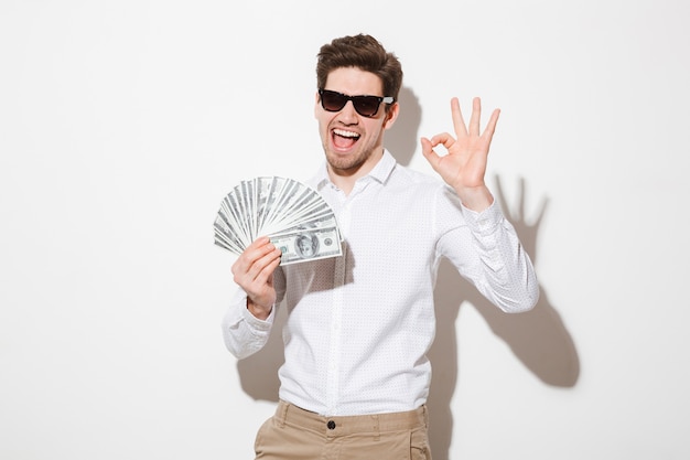 Фото счастливый победитель человек в рубашке и солнцезащитные очки, улыбаясь, держа веер денег в долларовых банкнотах и показывая хорошо символ, изолированных на белой стене с тенью