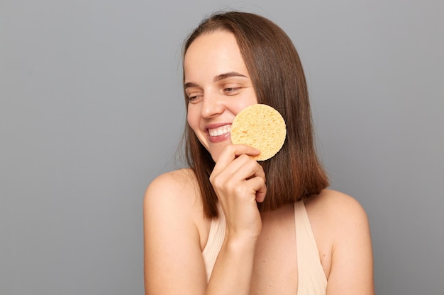 Photo of happy young adult girl with gentle smile holding cosmetic sponge near face isolated on gray background looking away enjoying cosmetic procedures