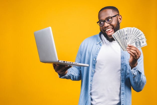 Фото счастливого молодого афро американского красивого человека представляя используя портативный компьютер держа деньги.