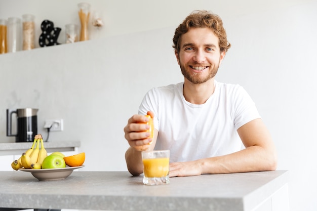 Photo of a happy young bearded man at the table at home make a juice with fruits.