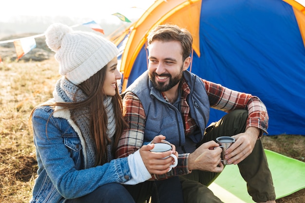 熱いお茶を飲みながら山を越えてキャンプする無料の代替休暇でテントを持って外で幸せな若い愛情のあるカップルの写真。