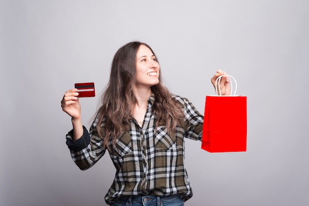 Photo of happy young woman holding credit card and red shopping bag and looking away