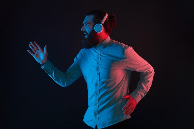 Photo of hipster man with beard wearing wireless headphones and running over dark background with neon light