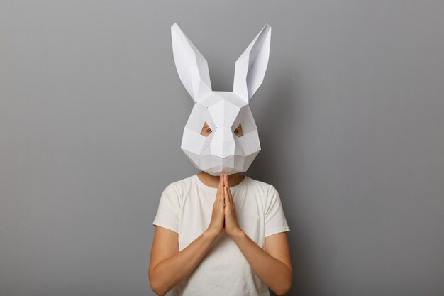 Photo of hopeful woman wearing white Tshirt and paper rabbit mask standing with palms together isolated over gray background praying or pleading asking to forgive