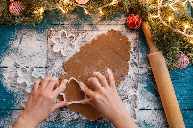 Photo of human hands, rolling pin, dough, spruce branches, garlands, biscuit molds on blue table