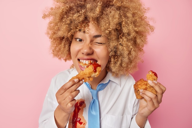 Photo of hungry young european woman eats delicious fried
chicken nuggets with ketchup winks eye enjoys eating fatty
appetizing unhealthy food dressed in formal clothes isolated over
pink background