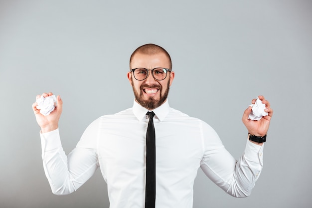 Photo of irritated angry businessman in white shirt and eyeglasses crumpling paper documents in both hands, isolated over gray wall