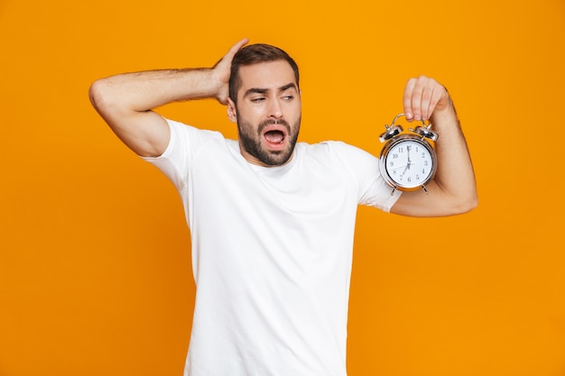 Photo of irritated man 30s in casual wear holding alarm clock, isolated
