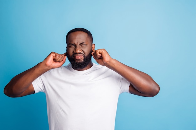 Photo of irritated man fingers close ears look side empty space wear white t-shirt over blue background