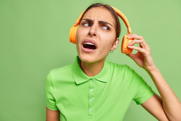 Photo of irritated woman takes off headphones and tries to hear something listens music with loud sound removes stereo accesory says what did you say I dont hear you poses against green wall