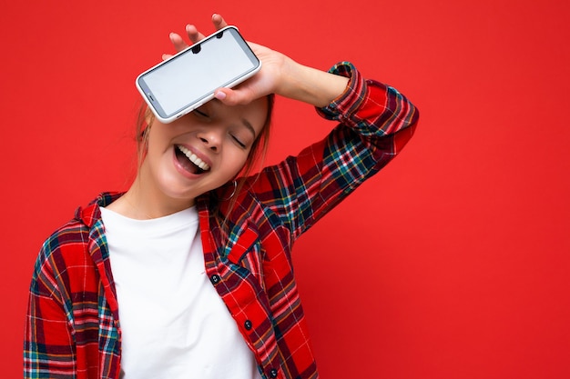 Фотография радостной красивой позитивной молодой блондинки, стоящей изолированно над красной стеной в повседневной красной рубашке и белой футболке, показывающей мобильный телефон с пустым экраном для макета
