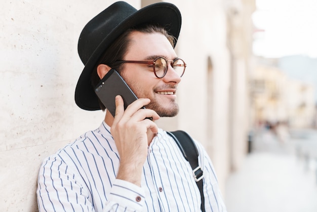 Photo of joyful caucasian man wearing eyeglasses and black hat talking on cellphone and smiling while leaning on wall in city street