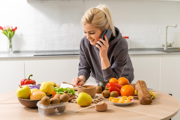 自宅のキッチンのインテリアで新鮮な野菜とサラダを調理しながらスマートフォンを持っているうれしそうな白人女性の写真。