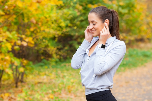 緑豊かな公園で休んでいる間、スポーツウェアでうれしそうなフィットネス女性30代のBluetoothイヤーポッドに触れて携帯電話を保持している写真