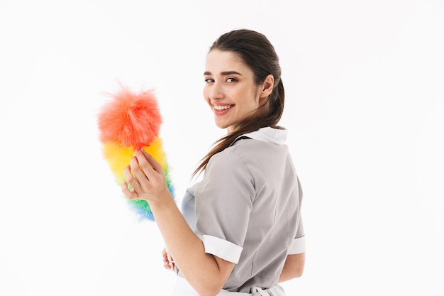 Foto di gioiosa cameriera 20s vestita in uniforme che tiene in mano uno spolverino colorato mentre fa i lavori di casa e pulisce la stanza isolata su un muro bianco