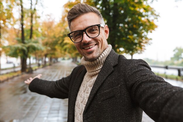 Foto foto di un uomo gioioso di 30 anni che indossa abiti caldi e sorride mentre cammina all'aperto attraverso il parco in autunno