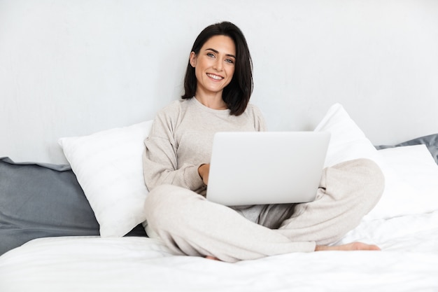 Фотография радостной женщины 30 лет с ноутбуком, сидя в постели с белым бельем в уютной квартире