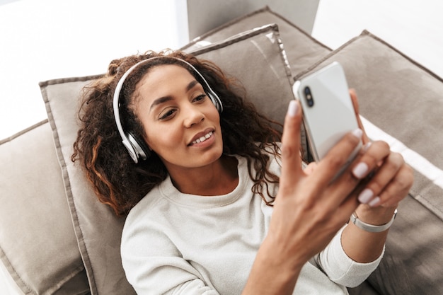 Foto foto di gioiosa donna afro-americana che indossa le cuffie tenendo il telefono cellulare, mentre giaceva sul divano in un luminoso appartamento