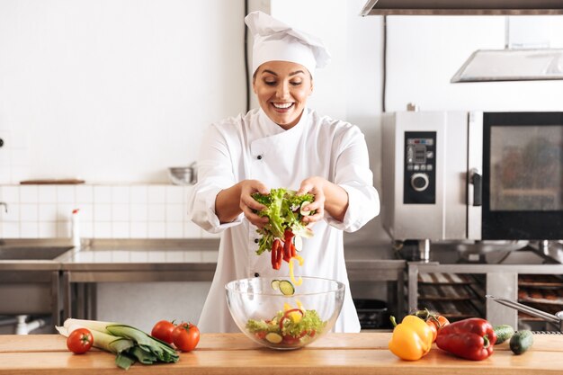 レストランのキッチンで、新鮮な野菜のサラダを作る白い制服を着た楽しい女性シェフの写真