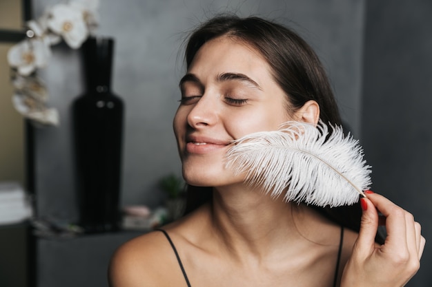 Photo of joyous woman with long dark hair and clean skin touching her face with big feather