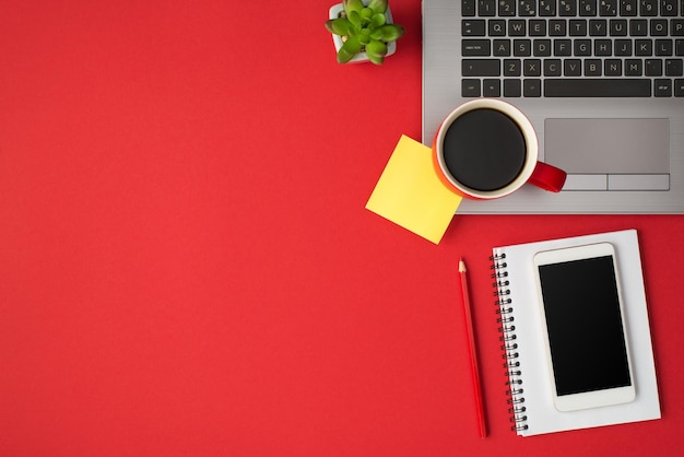 Above photo of laptop plant cup of coffee yellow sticker notepad phone and pencil isolated on the red backdrop with empty space