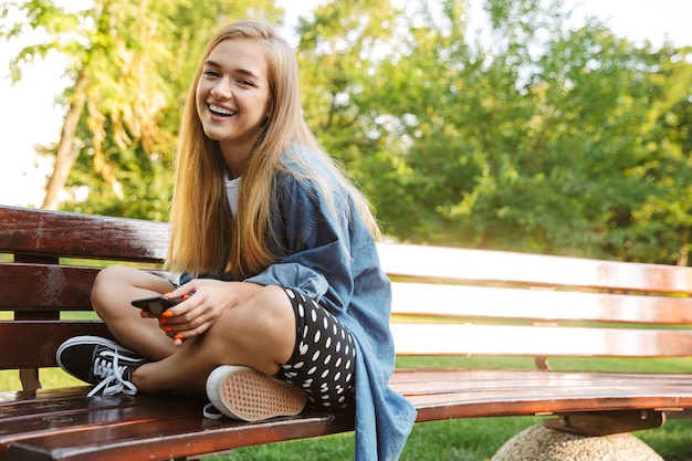 Photo of a laughing cheery emotional young girl outside in nature green park sit on a bench using mobile phone.