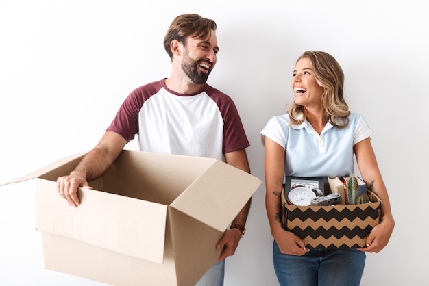 Photo of laughing couple in casual clothing holding cardboard boxes while looking at each other isolated over white wall