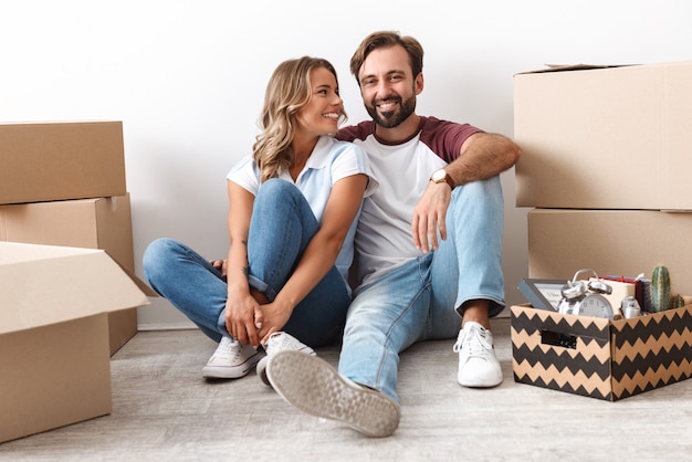 Photo of laughing couple in casual clothing hugging while sitting near cardboard boxes isolated over white wall