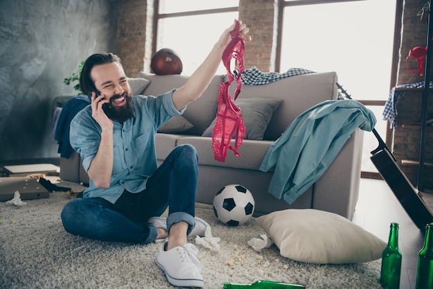 Photo of laughing hipster guy holding telephone telling friends\
intimacy details of his active naughty life night bad person watch\
red bra hands sitting floor trash after party flat indoors